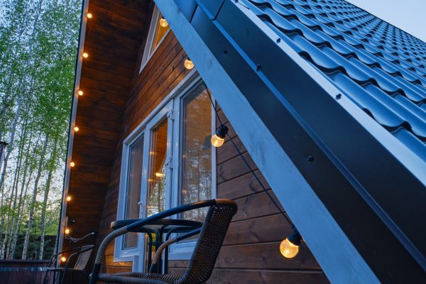 A charming wooden cabin is adorned with glowing string lights, creating a warm atmosphere. A single chair sits on the deck, inviting relaxation as twilight settles in among the trees.